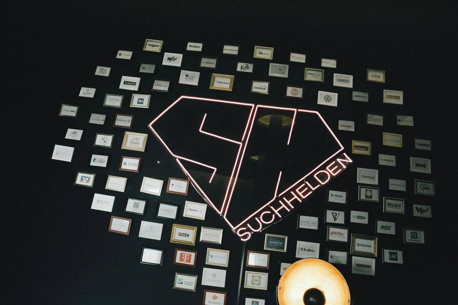 Kundenwand mit gerahmten Logos rund um das SH-Neonlogo im Suchhelden-Büro in Osnabrück