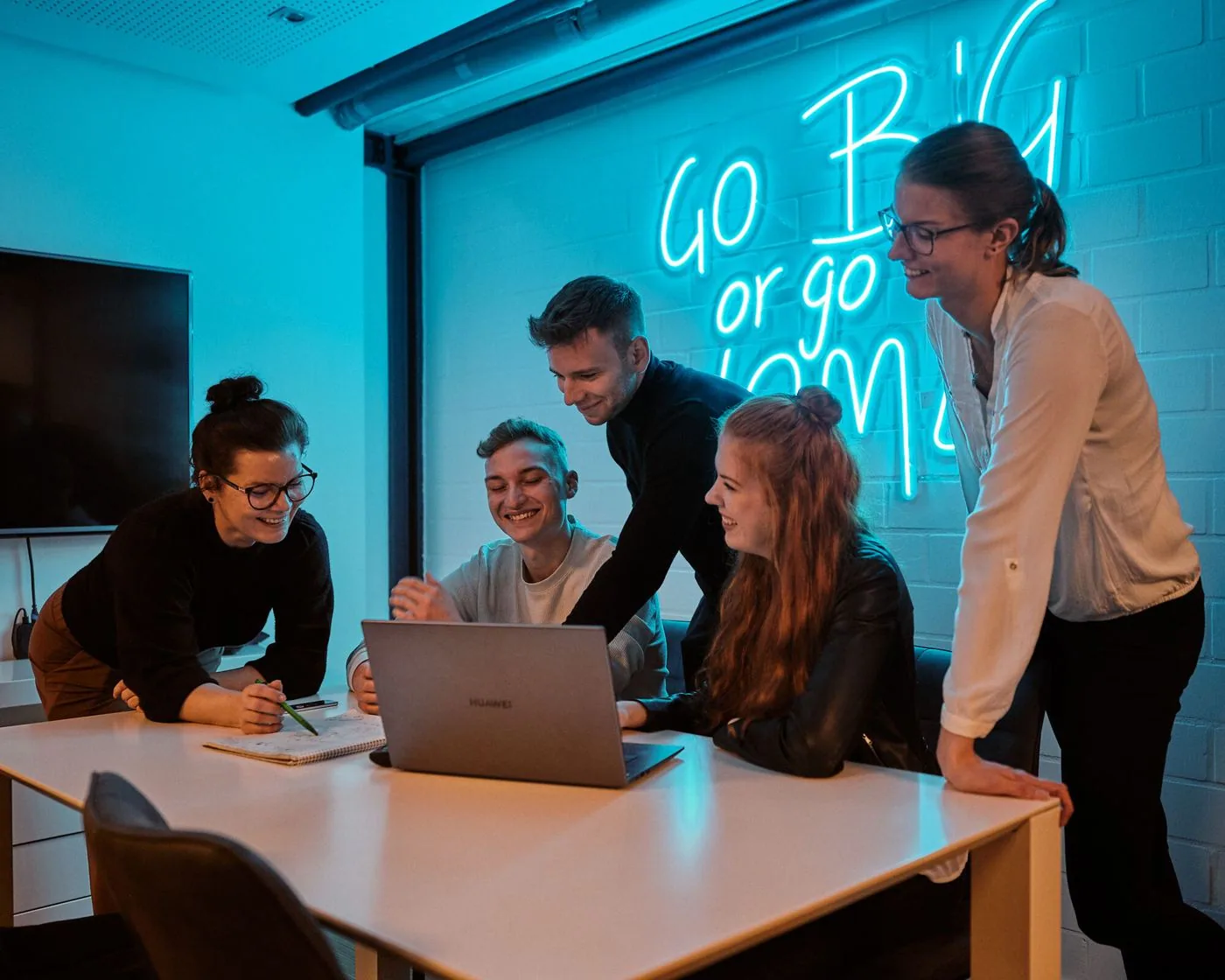 Suchhelden-Team bespricht ein Projekt am Laptop vor dem Neonschriftzug „Go big or go home“ in Osnabrück